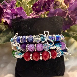 May include: A set of three stacked bracelets featuring faceted beads in shades of blue, purple, and red. Each bracelet is accented with silver-toned spacers and a small silver-toned bow charm. The beads vary in texture, including smooth, crackled, and translucent finishes, creating a layered look.