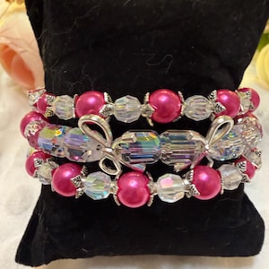 May include: A multi-strand bracelet featuring alternating hot pink round beads and clear faceted crystal beads. The design includes silver-toned metal spacers and a central silver-toned bow charm, all arranged on a black velvet display pillow.