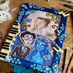 May include: A framed artwork featuring characters from the animated film "Corpse Bride." The blue frame is decorated with butterflies and flowers. The artwork depicts the characters in a colorful, cartoon style, with a piano key design on the frame.