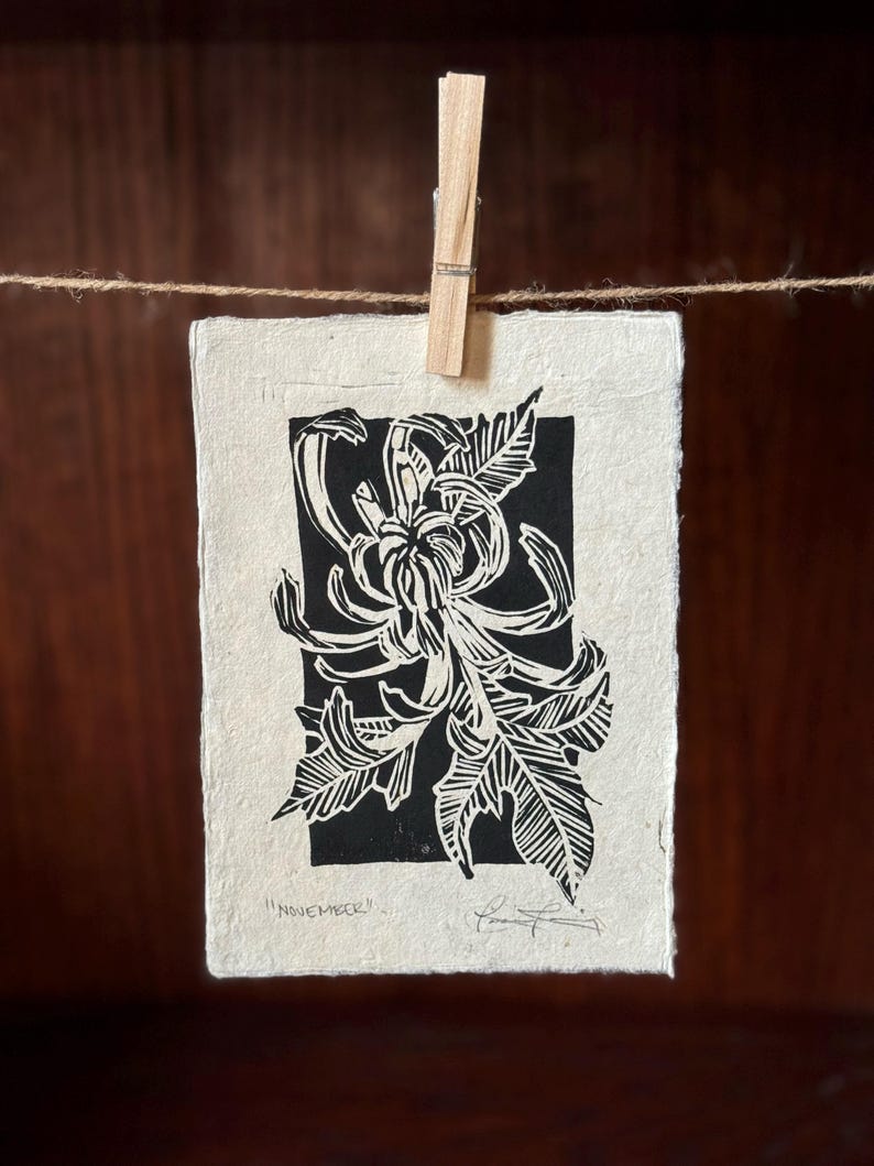 K&ouml;nnte beinhalten: Schwarz-wei&szlig;er Linolschnitt einer Chrysanthemenbl&uuml;te mit dem Text "November" unten.