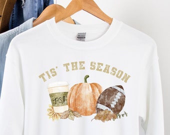 Puede incluir: Sudadera blanca con el texto dorado "TIS' THE SEASON". El diseño incluye una taza de café, una calabaza y un balón de fútbol americano. Un sombrero marrón y una camisa vaquera cuelgan detrás de la sudadera.