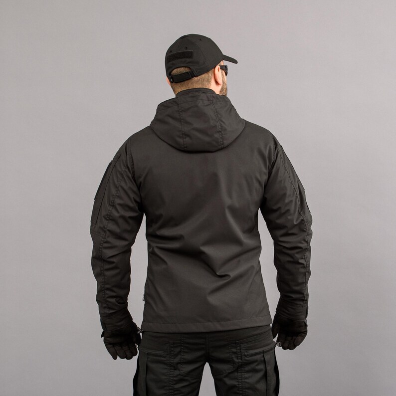 May include: Black hooded jacket with multiple pockets and a black baseball cap. The jacket has a zipper closure and is worn with black gloves and pants. The background is a solid gray.