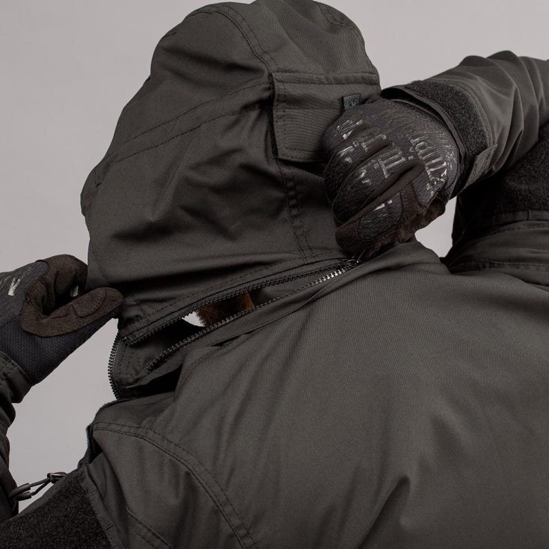 May include: Black hooded jacket with a zipper closure. The jacket has a pocket on the hood and is being zipped up by a person wearing black gloves. The jacket appears to be made of a durable, weather-resistant material.