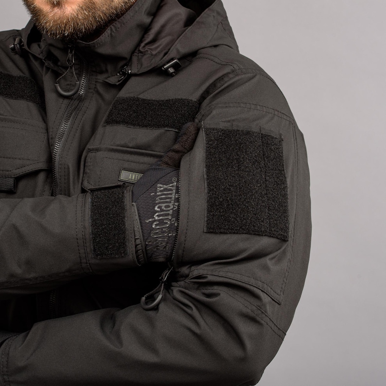 May include: Black tactical jacket with a hood and multiple pockets. The jacket features velcro patches on the sleeves and chest. The gloves have the word "Mechanix" printed on them.