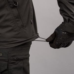 May include: Close-up of dark gray tactical pants and jacket. The jacket features a drawstring at the waist and a small pocket. A gloved hand is pulling the drawstring. The pants have a zippered pocket and a cargo pocket.