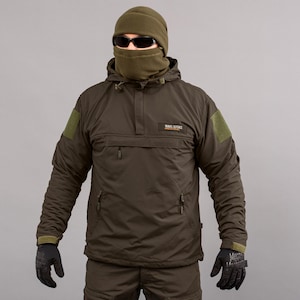 Könnte beinhalten: Olivgrüne Anorakjacke mit Kapuze, Vordertasche und dem Text "TRAVEL DEFENCE". Die Jacke hat eine fleecegefütterte Sturmhaube und Sonnenbrille. Die Person trägt schwarze Handschuhe.