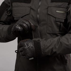 May include: Black tactical jacket with multiple pockets and a zipper closure. The jacket is paired with black gloves featuring the text "Mechanix Wear". The jacket has the word "ANTITERROR" on a small label.