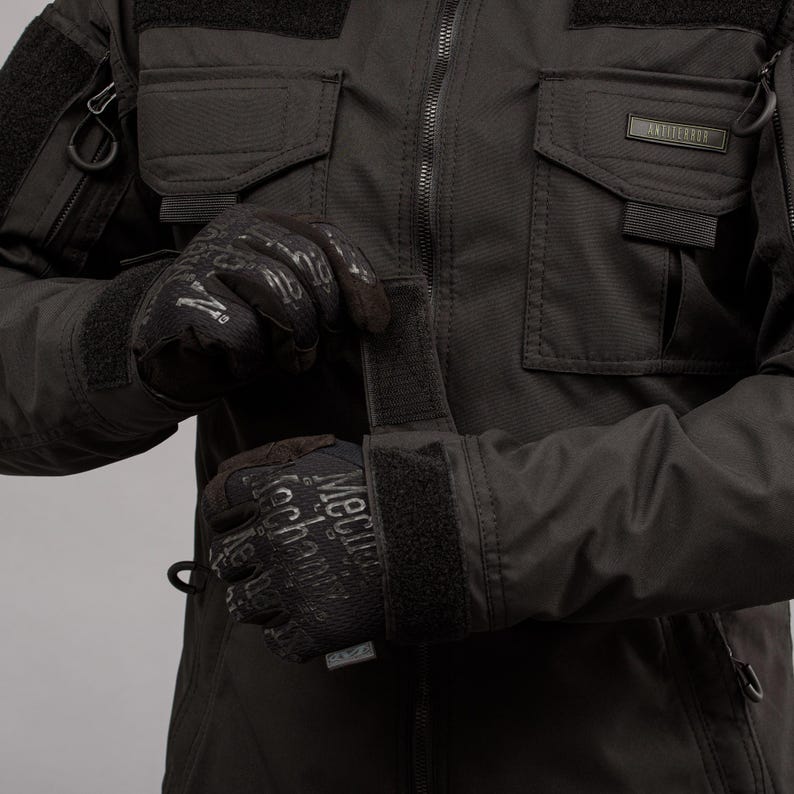 May include: Black tactical jacket with multiple pockets and a zipper closure. The jacket is paired with black gloves featuring the text "Mechanix Wear". The jacket has the word "ANTITERROR" on a small label.