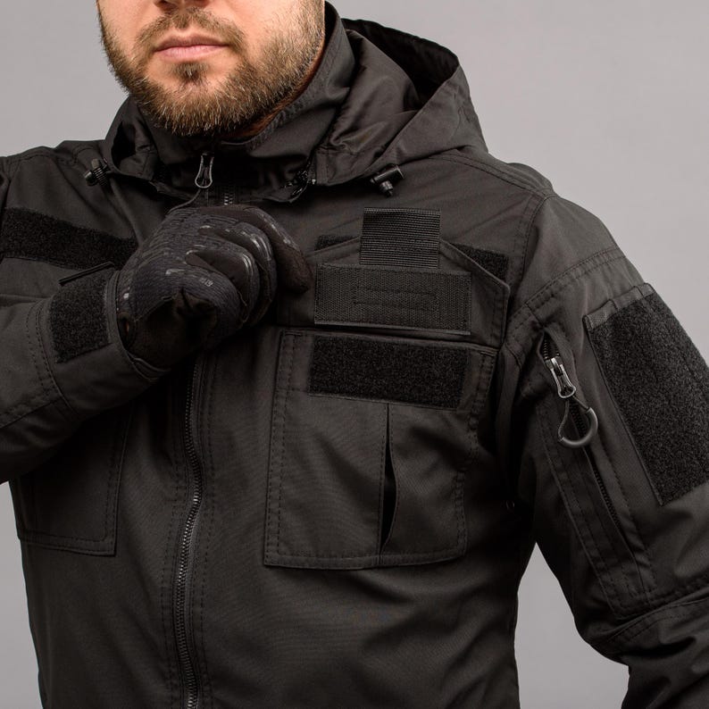 May include: Black tactical jacket with a hood, multiple pockets, and Velcro patches. The jacket features a zipper closure and a small pocket on the sleeve. The person in the image is wearing black gloves.