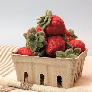 May include: A basket filled with red strawberries with green leaves. The strawberries are in a brown paper container, resting on a cream-colored towel with brown stripes. The background is a light gray.