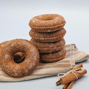 May include: A stack of five brown donuts dusted with white powder, next to a single donut and a bundle of cinnamon sticks tied with twine. The donuts rest on a striped beige and white cloth, all against a light blue background.