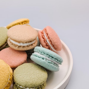 May include: A collection of colorful macarons in shades of pink, green, yellow, and beige, arranged on a white plate. Each macaron has a creamy white filling, creating a visually appealing dessert display.