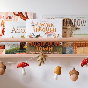 May include: A wooden shelf with a selection of children's books, including "The Very Lazy Elephant", "Because Acorn", "A Walk Through", and "Mushroom Town". A felt garland with mushrooms, acorns, and leaves hangs below the shelf.