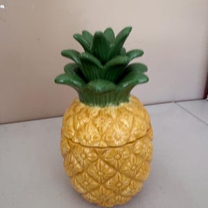 May include: A ceramic pineapple-shaped container with a lid. The container is yellow with a textured surface, and the top is green. The pineapple-shaped container is a decorative item.