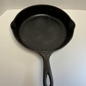 May include: A black cast iron frying pan with a long handle and a rounded cooking surface. The number "8" is embossed on the handle. The pan has a well-seasoned, matte finish, and is designed for stovetop or oven use.