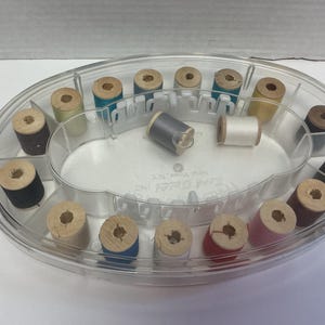 May include: A clear plastic oval sewing kit containing wooden spools of thread in various colors. The spools are arranged in a divided tray. Some spools are black, blue, pink, and yellow.