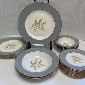 May include: A set of ceramic dinnerware featuring a cream-coloured centre with a floral motif of green leaves and brown blossoms, surrounded by a wide slate grey rim and a thin gold-tone edge. The collection includes round plates and bowls.