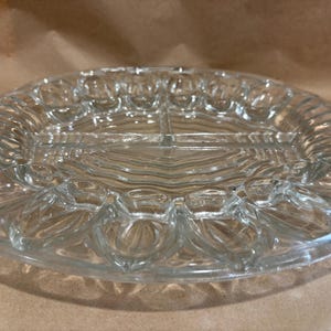 May include: Clear glass serving platter with a divided design. The oval-shaped platter features a textured, ribbed pattern around the edges and a central section divided into three compartments. The clear glass allows for visibility of the food.