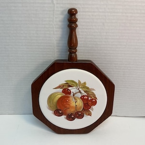 Pode incluir: Trivet de madeira com uma telha de cerâmica com um desenho de frutas. O trivet tem uma moldura e uma alça de madeira marrom escura. A telha exibe uma ilustração colorida de pêssegos e cerejas.