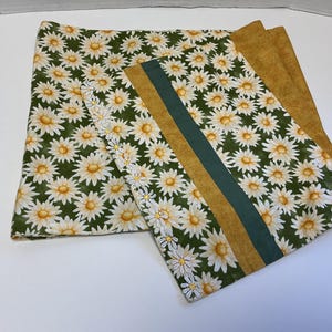 May include: Two pieces of fabric with a daisy pattern. One is a large rectangle, the other is a smaller rectangle with a green stripe and a daisy border. The background is a dark green with white and yellow daisies.