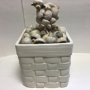 May include: A white ceramic basket with a woven pattern, filled with white ceramic mushrooms. A white ceramic sheep figurine stands on top of the mushrooms.