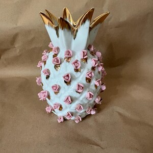 Puede incluir: Un jarrón de cerámica con forma de piña blanca que presenta detalles pintados en oro en las hojas superiores y numerosas rosas pequeñas de color rosa claro en relieve aplicadas sobre el cuerpo, creando una superficie floral texturizada.