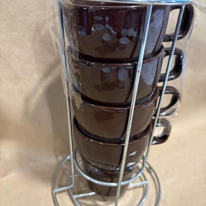 May include: A set of four brown ceramic coffee cups with handles, stacked in a silver metal rack. The cups are glossy and appear to be new, still in their original packaging. The rack has a circular base and a handle for easy carrying.