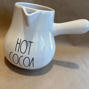 Puede incluir: Servidor de chocolate caliente de cerámica blanca con asa y boquilla. Las palabras "HOT COCOA" están escritas en letras negras y estilizadas en la parte delantera. El servidor está sobre una superficie de color neutro.