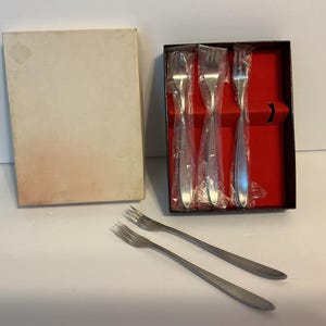 May include: A collection of stainless steel forks, some in plastic, displayed in a black box with a red interior. Two extra forks are outside the box. A beige cardboard container is also present.