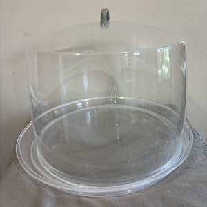 May include: A clear, round cake stand with a domed lid and a silver handle. The stand has a circular base and a clear, cylindrical cover. The stand is made of clear plastic and is designed to display and protect baked goods.