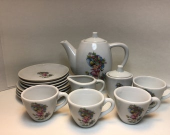 May include: A white porcelain children's tea set with a floral design featuring a girl in a hat. The set includes a teapot, creamer, sugar bowl, and four teacups and saucers.