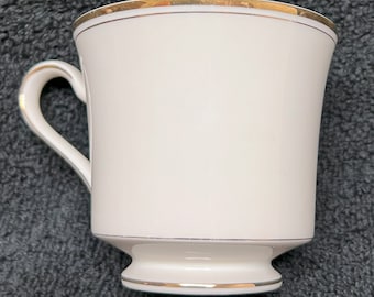 May include: A white ceramic teacup with a gold rim and a gold band around the base. The cup has a rounded shape and a handle.
