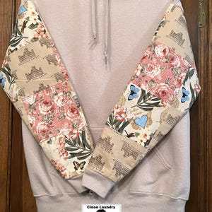 May include: A pale beige hoodie with patterned sleeves. The sleeves display a floral and butterfly design with the words "Downton Abbey". The hoodie has a front pocket and a drawstring hood. The bottom of the image has a logo that says "Clean Laundry Shirt Shop".
