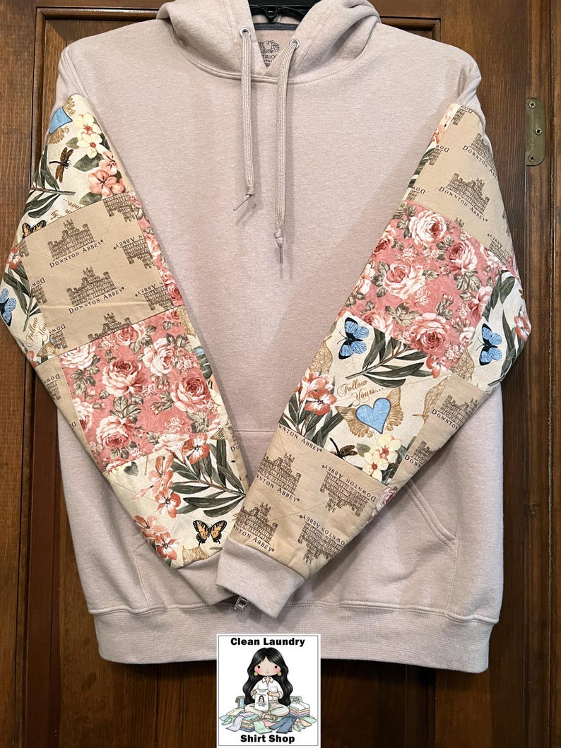 May include: A pale beige hoodie with patterned sleeves. The sleeves display a floral and butterfly design with the words "Downton Abbey". The hoodie has a front pocket and a drawstring hood. The bottom of the image has a logo that says "Clean Laundry Shirt Shop".