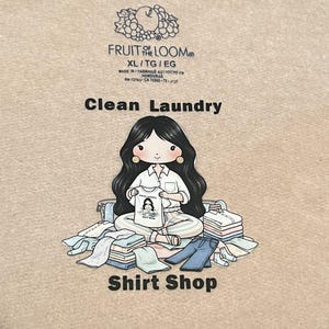 May include: A beige Fruit of the Loom XL t-shirt with the text "Clean Laundry" and "Shirt Shop". The shirt features a cartoon illustration of a woman holding a shirt with a graphic. The shirt is surrounded by folded clothes.
