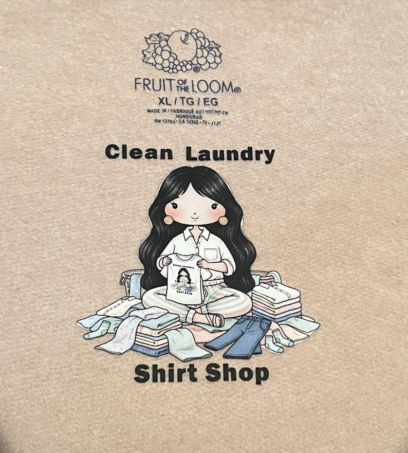 May include: A beige Fruit of the Loom XL t-shirt with the text "Clean Laundry" and "Shirt Shop". The shirt features a cartoon illustration of a woman holding a shirt with a graphic. The shirt is surrounded by folded clothes.