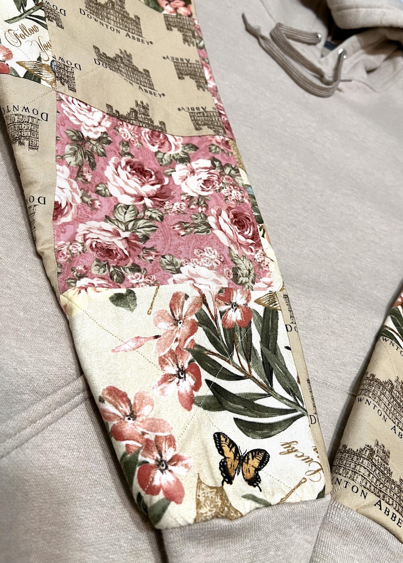 May include: Detail of a beige sweatshirt sleeve with a patchwork design. The patchwork includes floral patterns in pink and cream, a Downton Abbey print, and a butterfly. The sleeve is quilted.