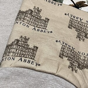 May include: Close-up of a light beige fabric featuring a repeating pattern of a brown building and the text "Downton Abbey". The fabric is sewn onto a grey textile.