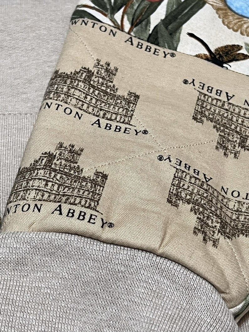 May include: Close-up of a light beige fabric featuring a repeating pattern of a brown building and the text "Downton Abbey". The fabric is sewn onto a grey textile.