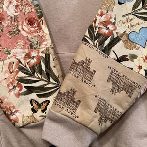 May include: A beige hoodie with quilted sleeves. The sleeves display floral and butterfly patterns, the text "DOWNTON ABBEY" and "Follow Yours...". The cuffs are ribbed and beige.