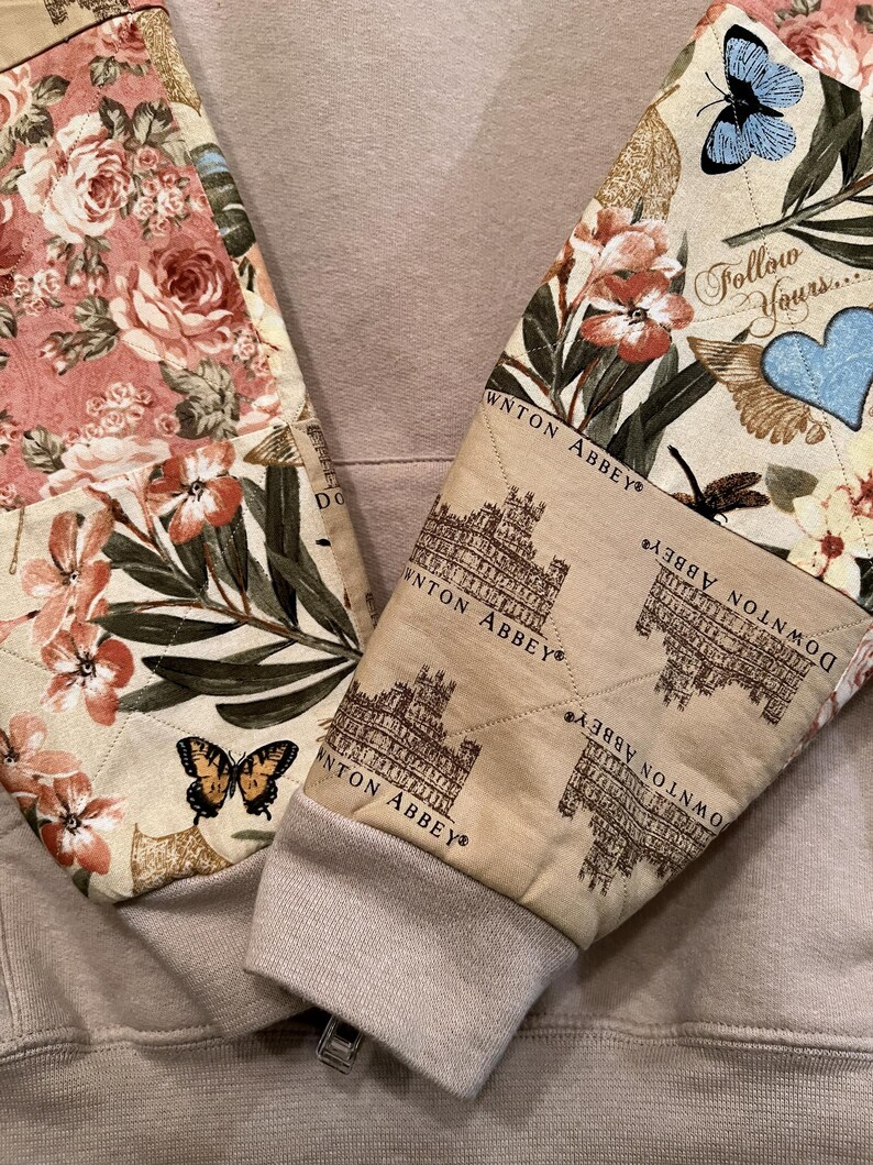 May include: A beige hoodie with quilted sleeves. The sleeves display floral and butterfly patterns, the text "DOWNTON ABBEY" and "Follow Yours...". The cuffs are ribbed and beige.