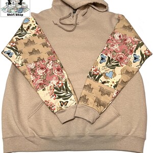 May include: A beige hoodie with patterned sleeves. The sleeves display a floral and butterfly design with the words "Downton Abbey". The hoodie has a drawstring hood and a front pocket.