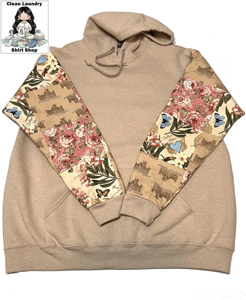 May include: A beige hoodie with patterned sleeves. The sleeves display a floral and butterfly design with the words "Downton Abbey". The hoodie has a drawstring hood and a front pocket.