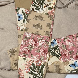May include: A beige hoodie with decorative patchwork panels. The patchwork includes floral designs in pink and cream, butterflies, and Downton Abbey text. The hoodie has a drawstring and a front pocket.
