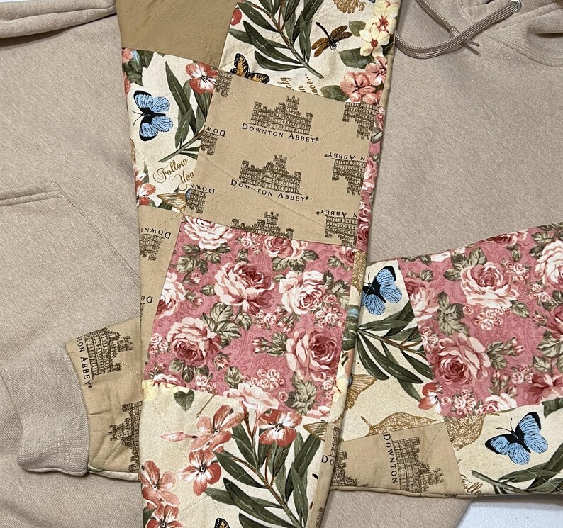 May include: A beige hoodie with decorative patchwork panels. The patchwork includes floral designs in pink and cream, butterflies, and Downton Abbey text. The hoodie has a drawstring and a front pocket.
