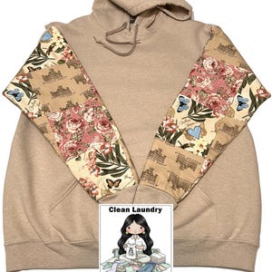 May include: A beige hoodie with patterned sleeves. The sleeves are decorated with a patchwork design featuring roses, butterflies, and text. The hoodie has a drawstring hood and a kangaroo pocket. The image includes a logo that says "Clean Laundry Shirt Shop."