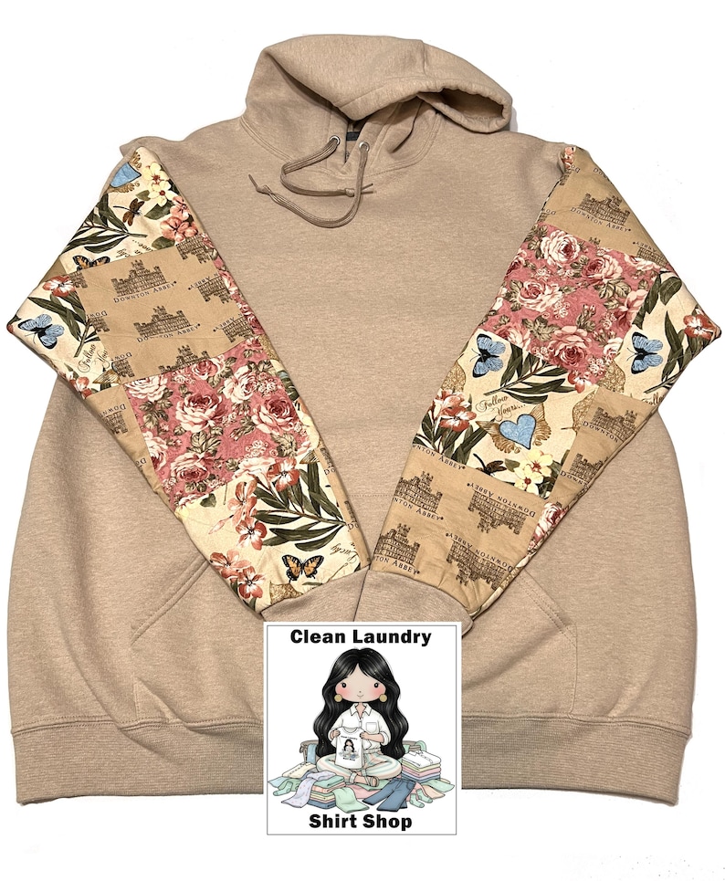 May include: A beige hoodie with patterned sleeves. The sleeves are decorated with a patchwork design featuring roses, butterflies, and text. The hoodie has a drawstring hood and a kangaroo pocket. The image includes a logo that says "Clean Laundry Shirt Shop."