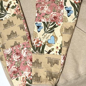 May include: Beige sweatpants with patchwork panels. The panels display Downton Abbey imagery, floral designs, and butterflies. Text on the panels reads "Follow Yours..." and "Downton Abbey". The cuffs are plain beige.