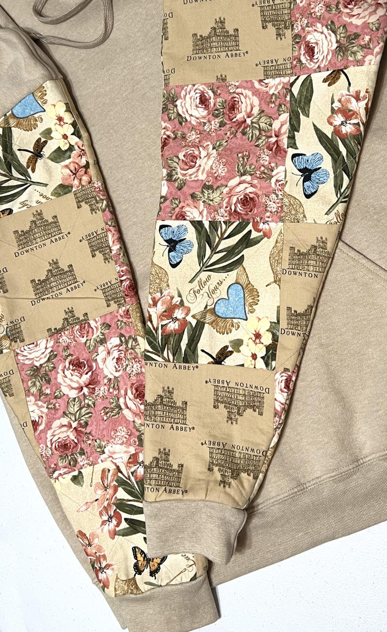 May include: Beige sweatpants with patchwork panels. The panels display Downton Abbey imagery, floral designs, and butterflies. Text on the panels reads "Follow Yours..." and "Downton Abbey". The cuffs are plain beige.