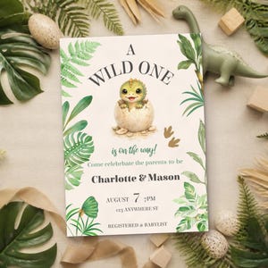 May include: A rectangular invitation card featuring a green baby dinosaur hatching from an egg, surrounded by tropical leaves. The text reads A WILD ONE is on the way! Come celebrate the parents to be Charlotte & Mason AUGUST 7 7PM 123 ANYWHERE ST REGISTERED AT BABYLIST.